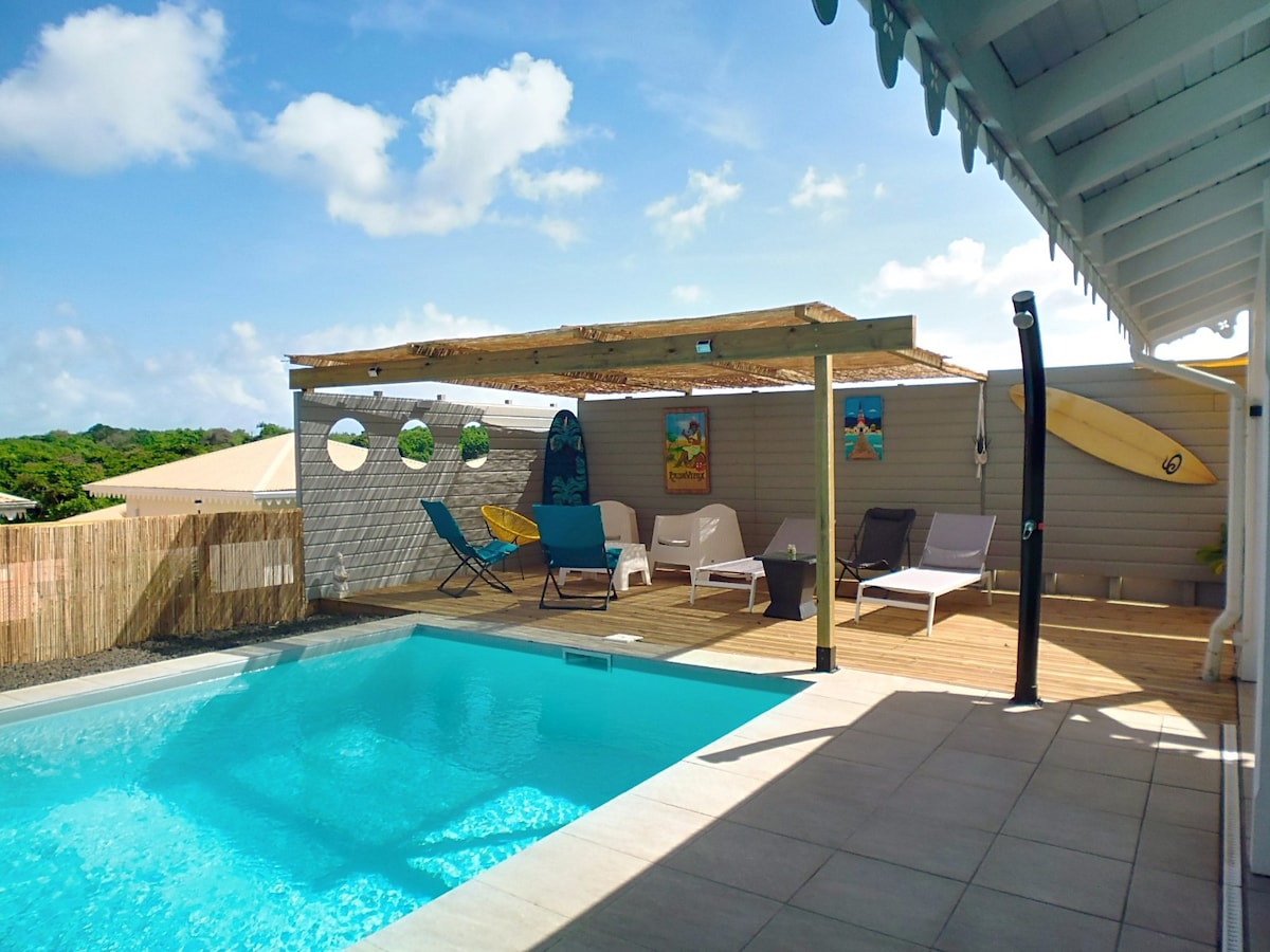 A pool area is shown, featuring a clear, inviting pool surrounded by a wooden deck. Several lounge chairs are positioned nearby, and a shaded seating area is highlighted. Decor includes surfboards and framed beach-themed artwork. The scene is complemented by a bright sky and greenery in the background.