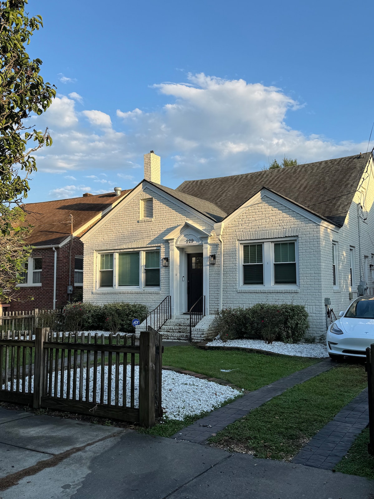 A quaint single-story home is depicted with a white brick exterior and a welcoming front porch. The property features a well-maintained lawn with white stones and a wooden fence. A driveway is visible, accommodating a parked vehicle, framed by lush greenery under a clear blue sky.