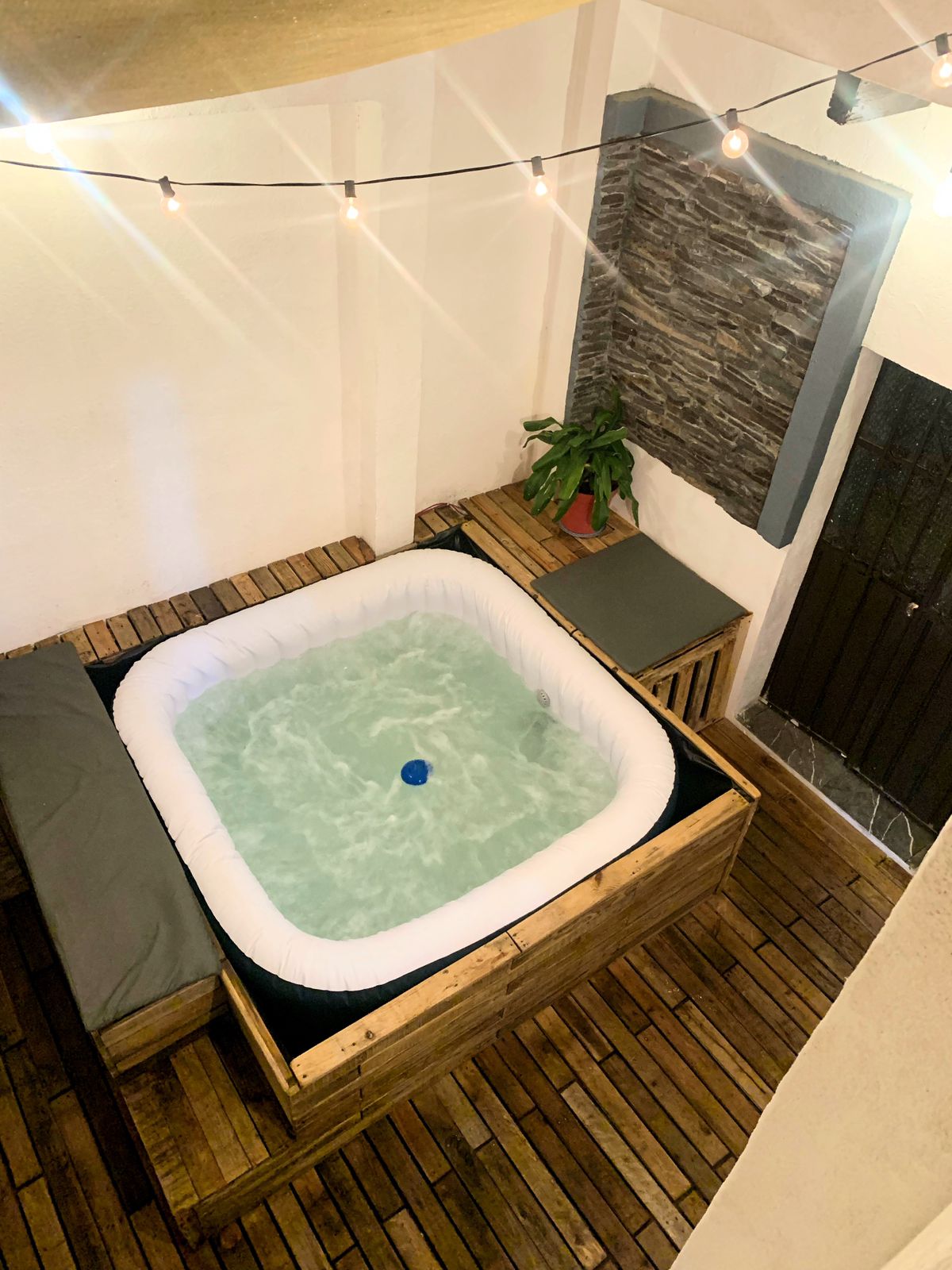 A cozy outdoor jacuzzi is situated within a wooden deck, illuminated by soft string lights overhead. Relaxing seating is positioned nearby, alongside a small green plant. The walls are adorned with natural stone accents, creating a serene environment for unwinding.