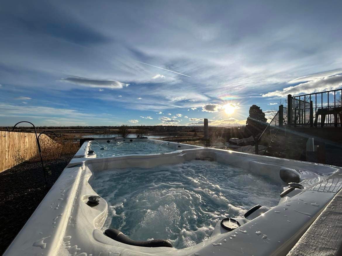 An 18-foot swim spa is showcased, with bubbling water reflecting the sunlight. In the background, a serene landscape is visible under a partially cloudy sky. The setting sun enhances the outdoor atmosphere, creating a tranquil environment for relaxation.