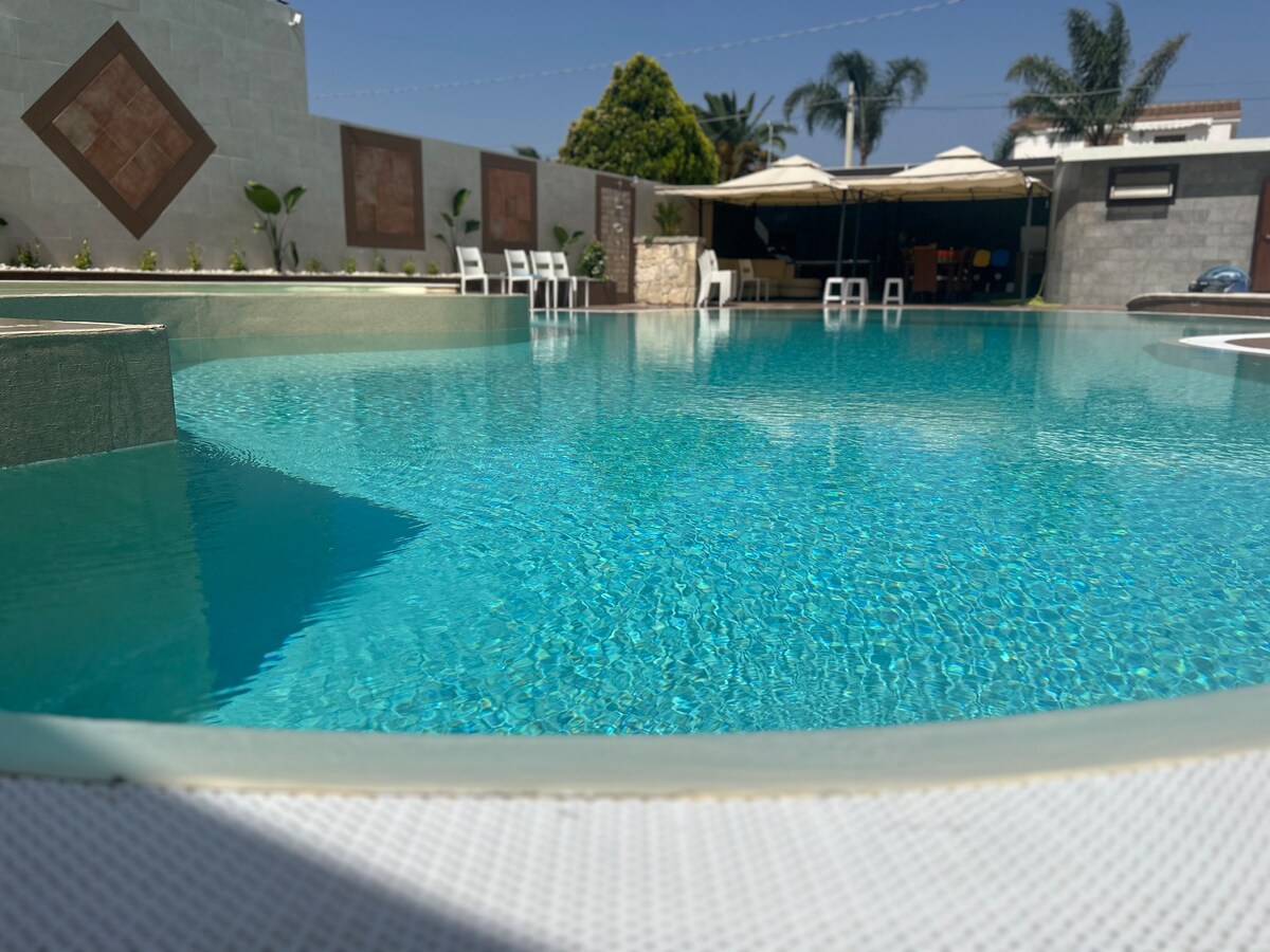 The shimmering water of the inviting pool reflects the clear sky, surrounded by a spacious patio area. White lounge chairs are positioned around the pool, complementing the lush greenery and decorative plants along the perimeter.