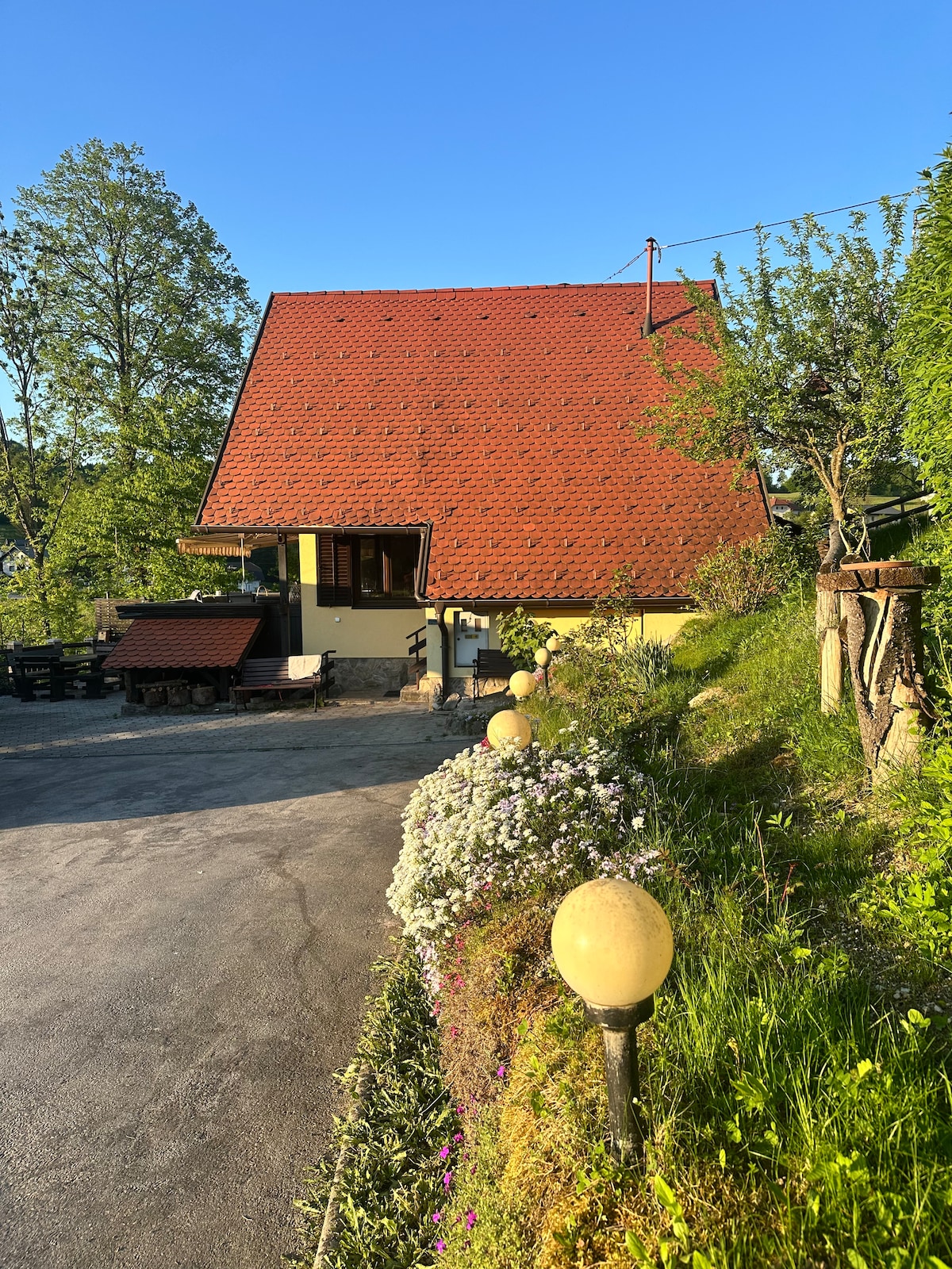 A charming house with a distinctive sloped roof is surrounded by greenery and colorful flowers. Soft sunlight bathes the exterior, highlighting the inviting entrance. Pathway lights line the drive, creating a welcoming ambiance as they lead to the home.