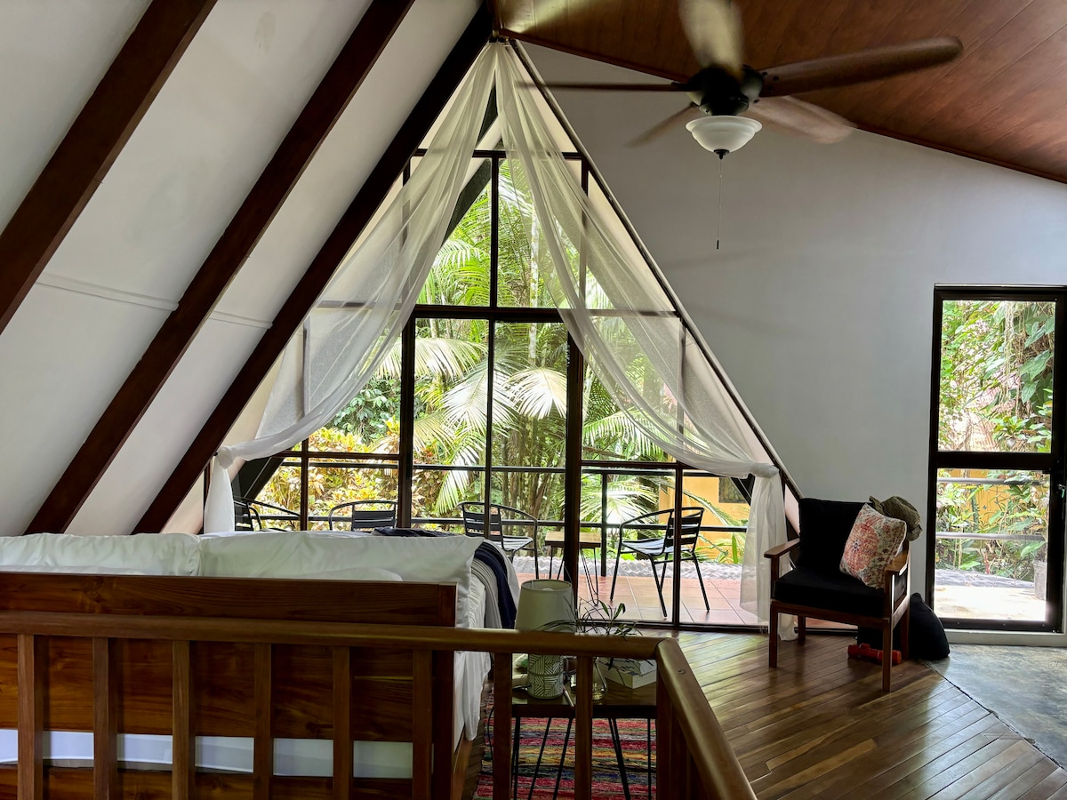 The interior features sloped wooden ceilings with large windows allowing natural light to flood the space. A spacious bed is accompanied by a small seating area and a decorative rug. Lush greenery is visible outside, enhancing the connection to nature.