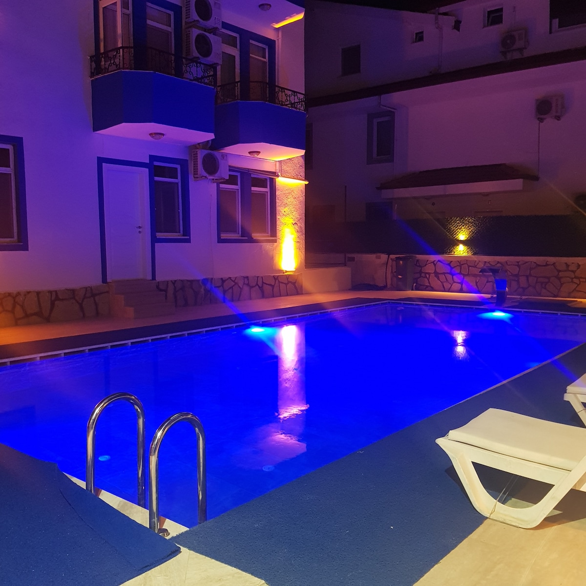 A nighttime view captures a pool area featuring illuminated blue water and surrounding lounge chairs. The vibrant lighting highlights the building's architecture and creates a tranquil atmosphere. The walls are accented with stone details, and air conditioning units are visible on the facade.