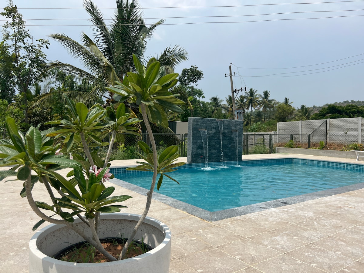 A serene swimming pool area is surrounded by tiled flooring and lush greenery. A stone wall with water features adds a soothing element. Tropical plants in circular planters enhance the natural setting, while distant palms gently sway in the background.