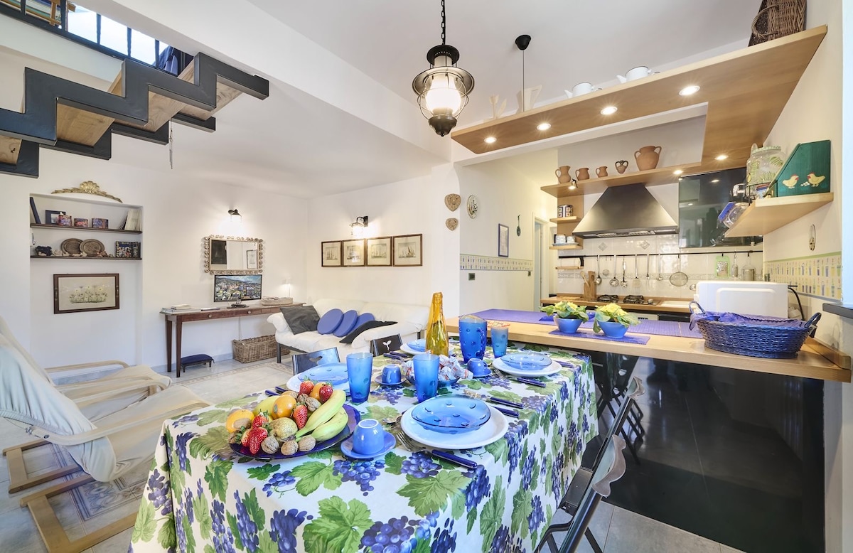 An open-plan living area features a dining table set with vibrant blue dishware and a floral tablecloth. A kitchen area is located in the background, equipped with modern appliances. A cozy seating area with a blue lounge chair and decorative items can be seen, along with a staircase leading to an upper level.
