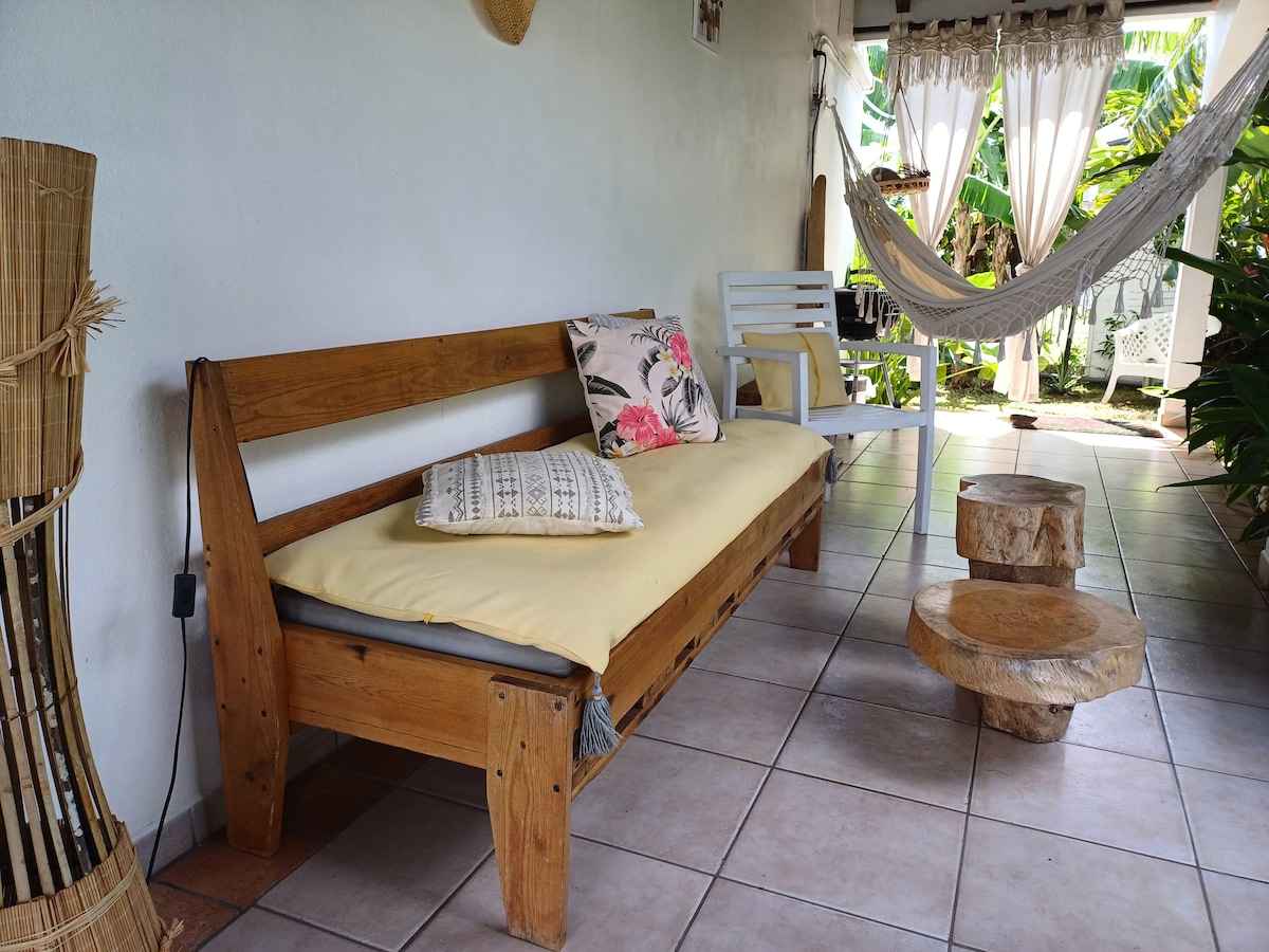 A relaxed seating area features a wooden bench with a yellow cushion and a decorative pillow, accompanied by a white chair and a wooden coffee table. A hammock is suspended nearby, with lush greenery visible in the background, creating a tranquil ambiance.