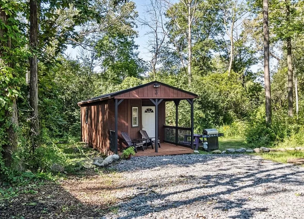 A rustic cabin is nestled among trees, featuring a front porch with two wooden chairs. A barbecue grill is positioned to the side, with a gravel pathway leading to the entrance. Natural light filters through the surrounding foliage, creating a serene outdoor setting.