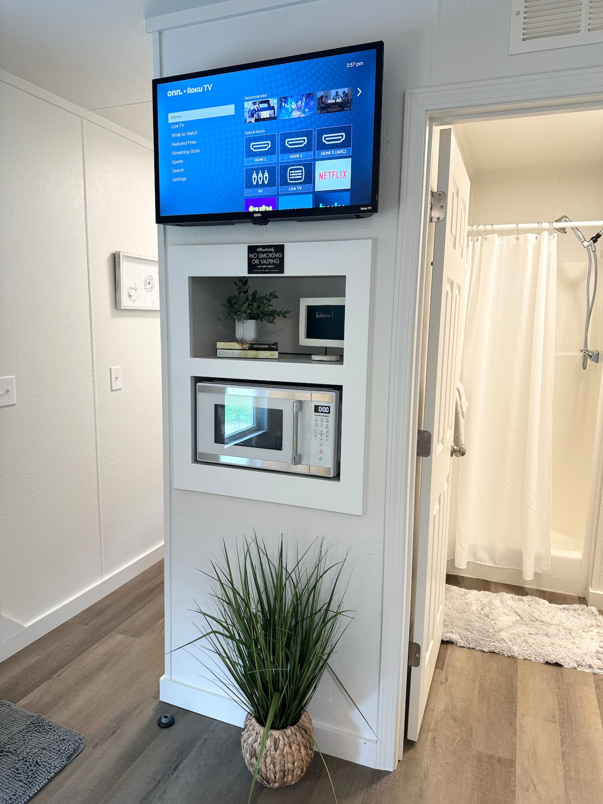 A modern nook features a mounted television displaying available streaming options above a neatly integrated microwave. A decorative plant in a woven basket adds a touch of greenery. The adjoining bathroom is partially visible, showcasing a shower area and light-colored curtain.