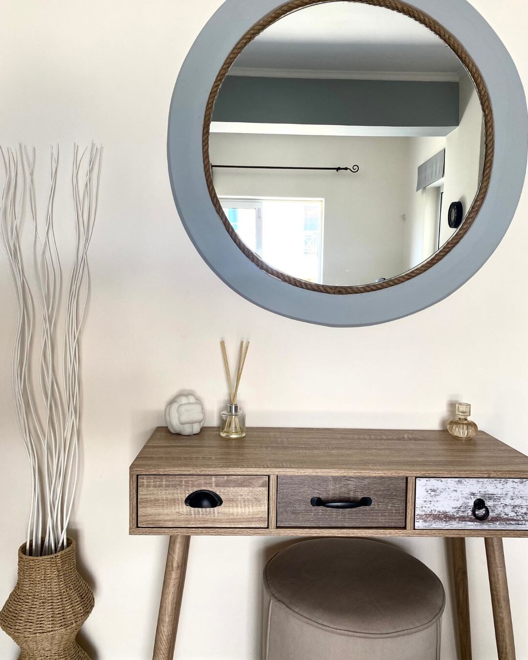 A decorative wooden vanity table with two drawers sits against a neutral wall. Above it, a round mirror with a rope border reflects the space. Next to the table, slender, white decorative branches are arranged in a tall vase.