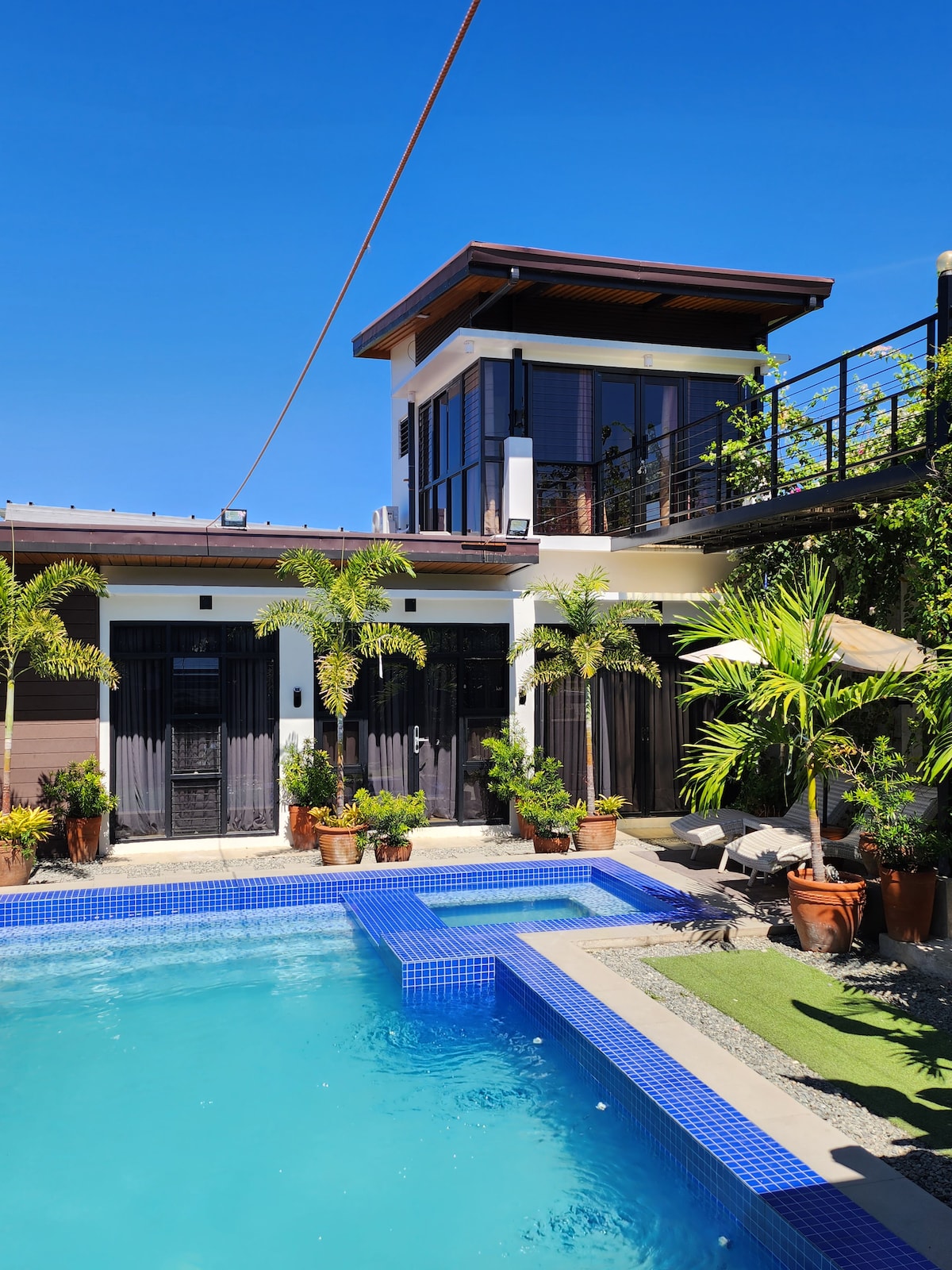 A refreshing swimming pool is framed by lush greenery and potted plants. The two-story guesthouse features dark-colored doors and large windows, providing ample natural light. A sun umbrella stands nearby, complementing the outdoor area under a clear blue sky.
