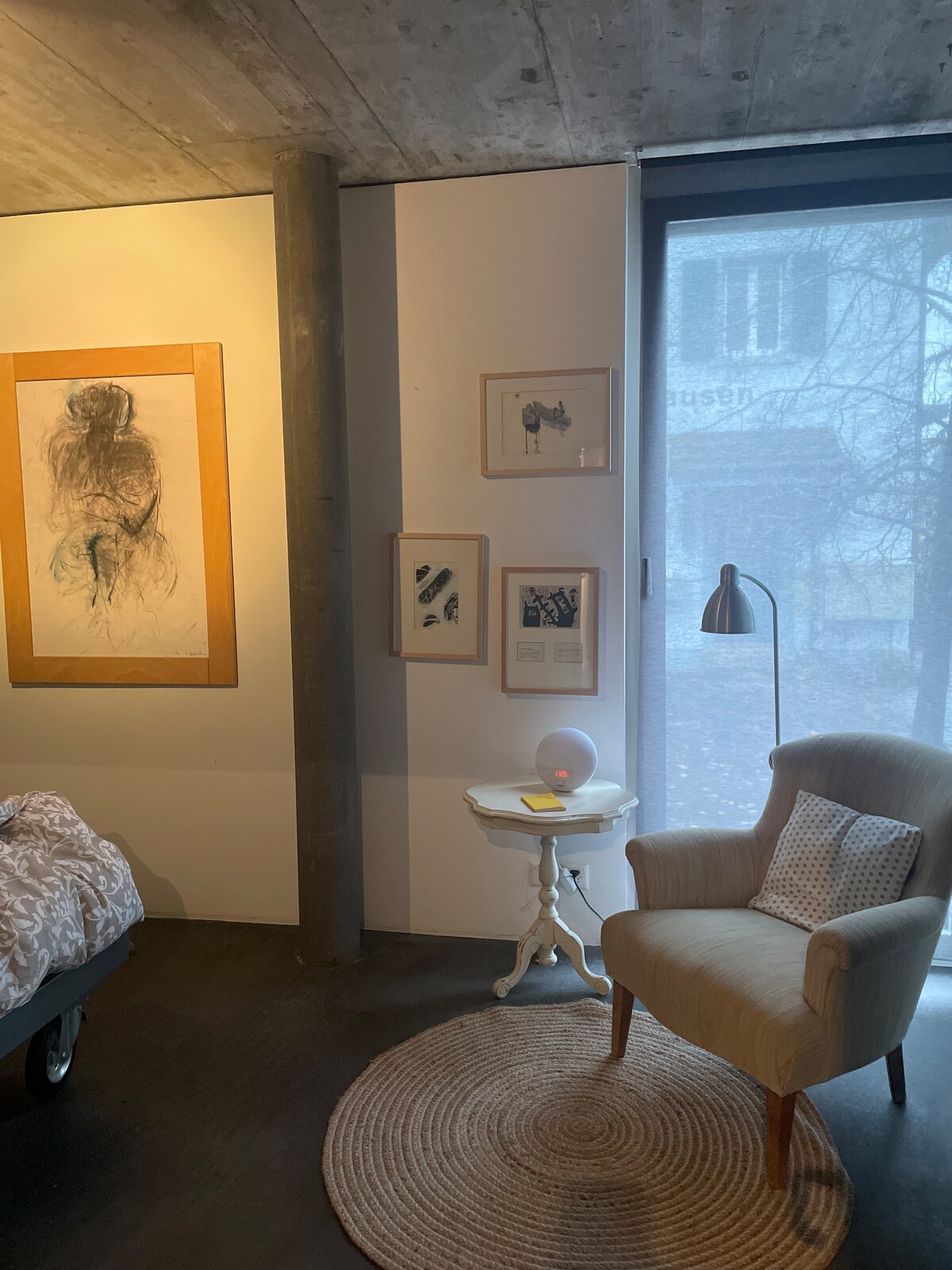 A cozy corner of a bedroom is seen, featuring a comfortable armchair next to a bedside table. Art pieces are displayed on the wall, and natural light filters through a large window, adding warmth to the space. A textured rug is placed underfoot.