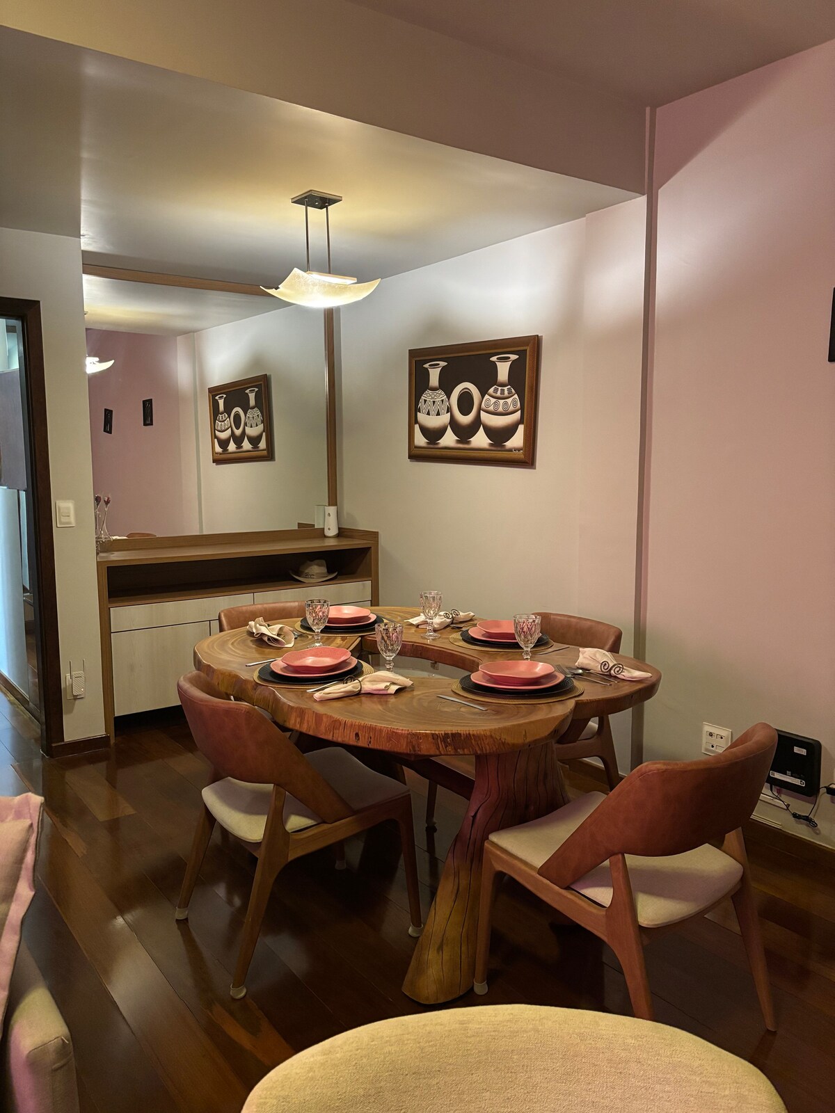 A dining area is showcased with a wooden table set for four, featuring elegant glassware and pink plates. Two framed artworks adorn the walls, and a light fixture hangs above, illuminating the space with a warm glow. The flooring is polished wood, contributing to a cozy ambiance.