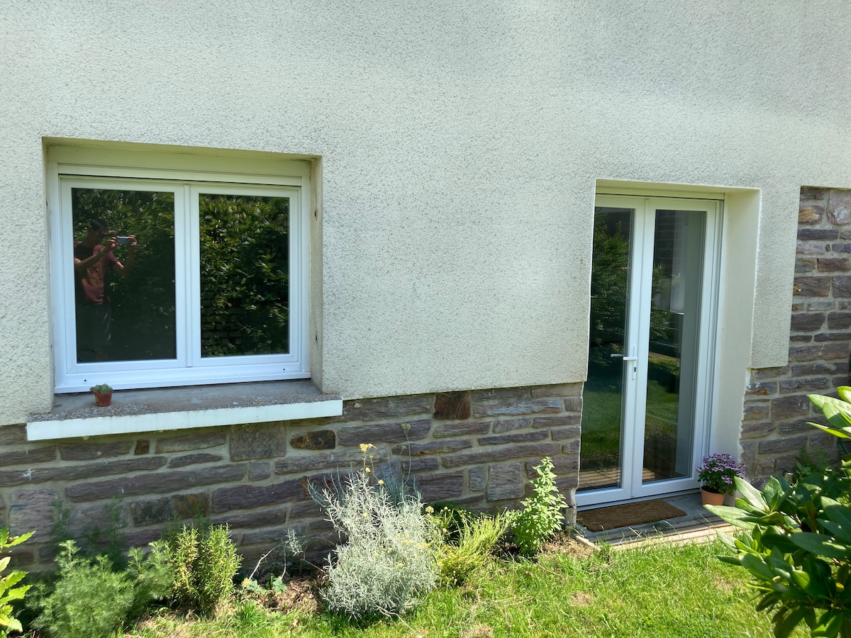 The exterior of the studio features a simple façade with a combination of light-colored walls and stone detailing. A door and a window are visible, allowing natural light to enter. Surrounding greenery adds a sense of tranquility to the entrance.