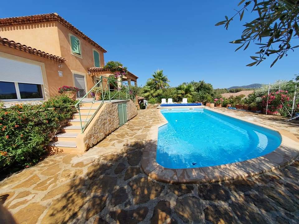A villa with a charming facade is set against a clear blue sky. A swimming pool is centrally located, surrounded by natural stone paving and lush greenery. Steps lead up to the entrance, flanked by flower beds that enhance the peaceful outdoor space.