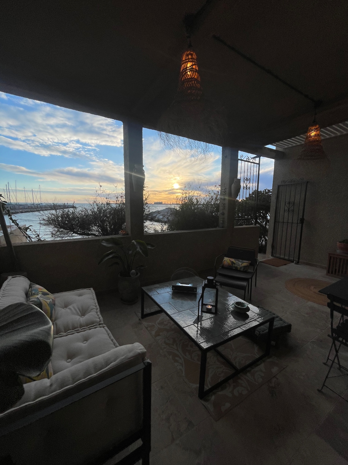 A cozy outdoor seating area features a comfortable sofa and a low table, surrounded by plants. Large windows allow for views of the sunset over the water, with boats visible in the distance. Soft lighting adds warmth to the tranquil space.