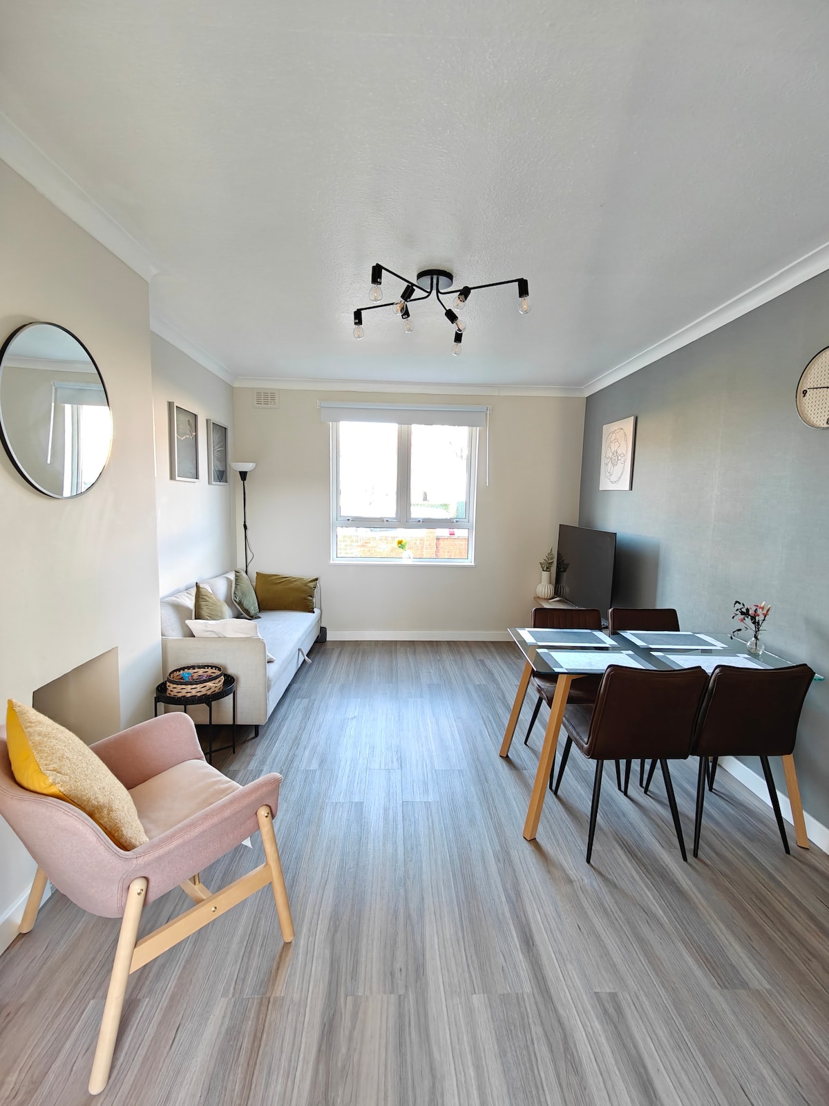 A bright living area features a combination of seating options, including a light-colored sofa and a pink accent chair. A dining table with four chairs is positioned beside a large window, allowing natural light to flood the space. Minimalist wall art decorates the walls.