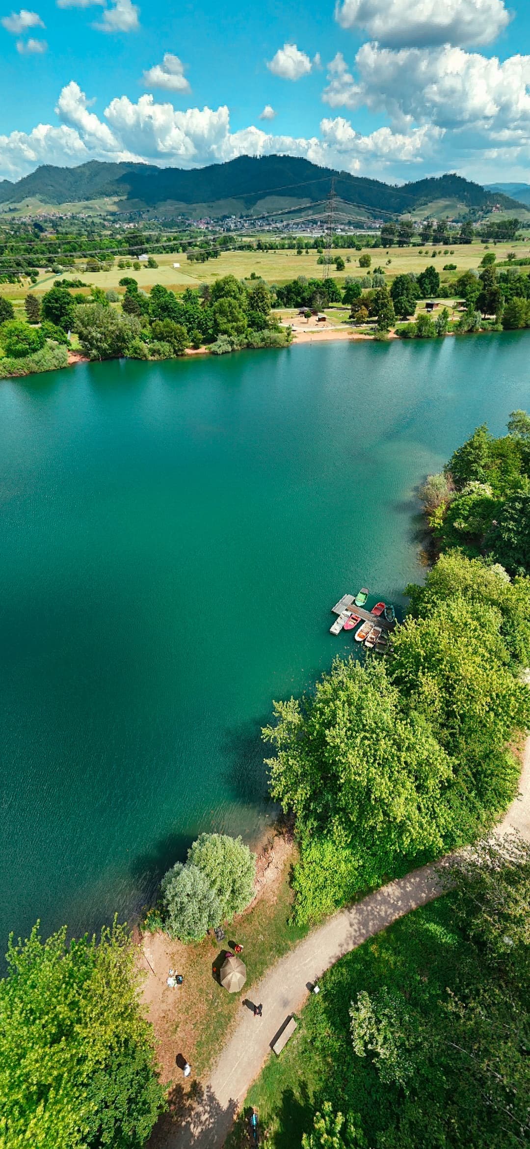 A panoramic view of a serene lake is captured, featuring calm turquoise water surrounded by lush greenery. The shoreline is lined with trees, and walking paths are visible. Small boats are docked near the water's edge, set against a backdrop of distant hills and clouds.