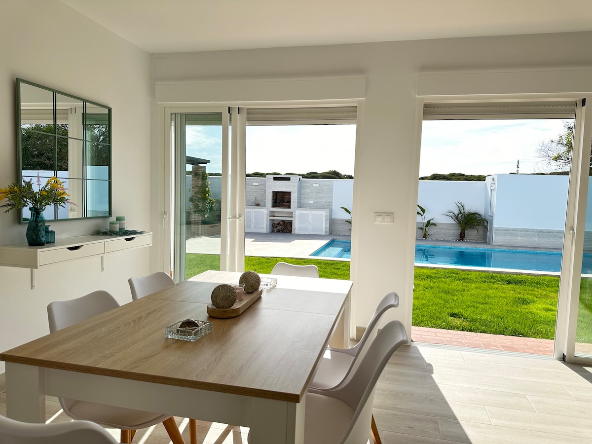 An open dining area is visible, featuring a light wooden table surrounded by white chairs. Glass sliding doors provide a view of the private pool and garden, allowing sunlight to brighten the space. A wall mirror and decorative items enhance the inviting atmosphere.