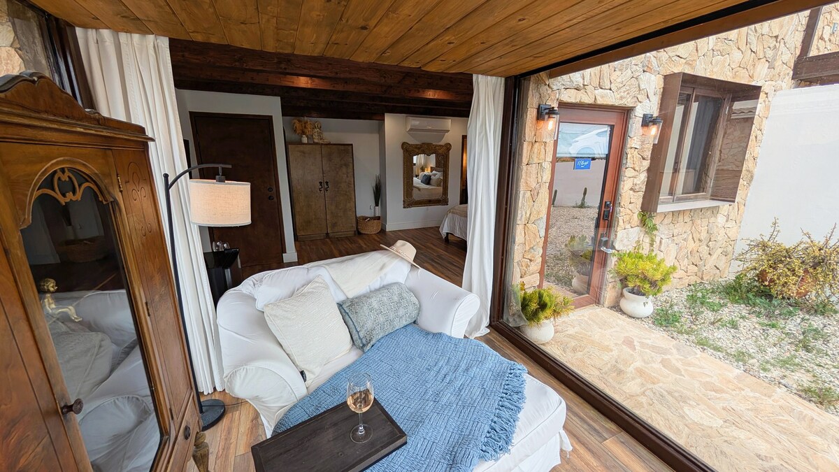 A cozy seating area features a white couch with a soft blue throw and a small wooden table holding a glass. Large glass doors open to an outdoor space, inviting natural light into the room. The decor includes a vintage wooden cabinet and a stylish lamp.