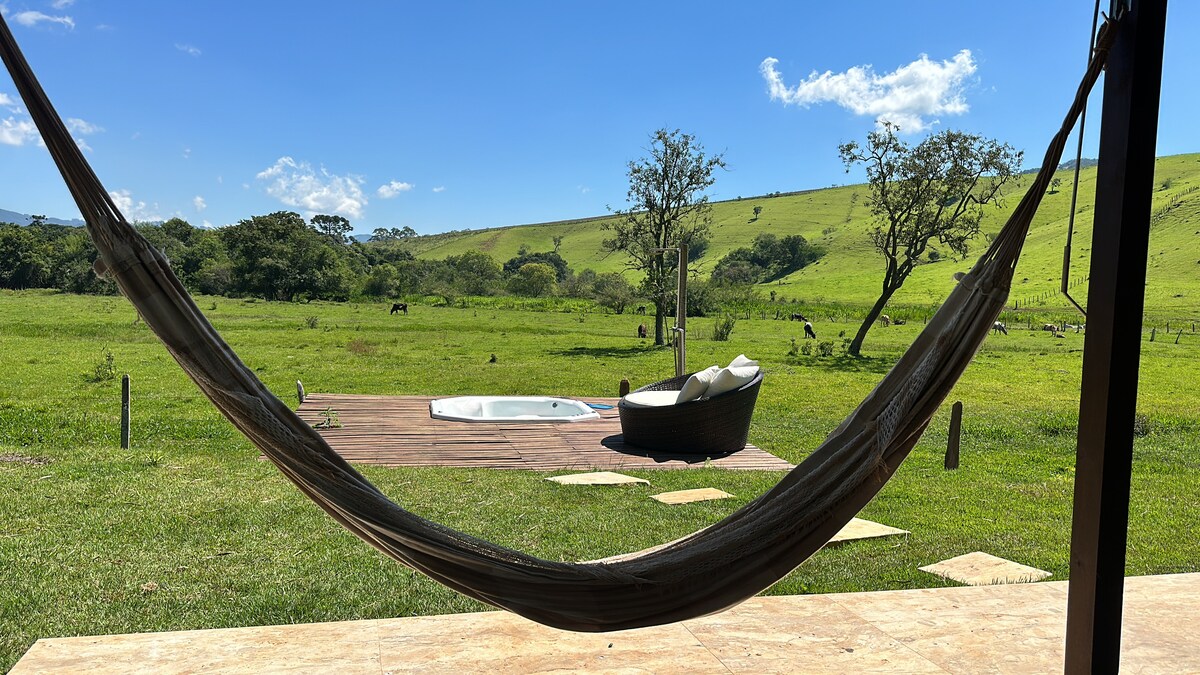 A hammock is suspended in front of a private outdoor area, offering a view of lush greenery and rolling hills. A cozy seating area is visible beside a hot tub, surrounded by manicured grass and the calming presence of trees in the distance.