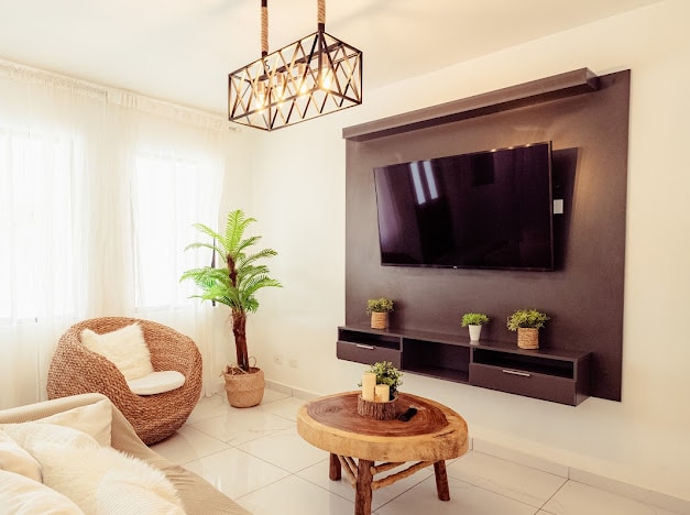 A modern living space features a flat-screen television mounted on a dark wall, complemented by two small potted plants. A round wooden coffee table sits in front of a comfortable armchair made of woven material. Natural light fills the room through sheer window treatments.