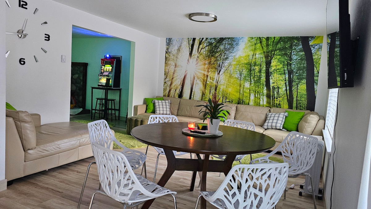 A spacious living area features a large circular table surrounded by modern white chairs. A sectional leather sofa offers ample seating, while a vibrant mural of a sunlit forest serves as an eye-catching backdrop. Natural light fills the space, enhancing the inviting atmosphere.