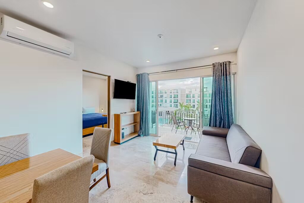 A bright living area features a gray sofa, a wooden coffee table, and a dining set with four chairs. Large glass doors open to a balcony with outdoor seating, allowing natural light to fill the space. A television is mounted on the wall opposite the sofa.