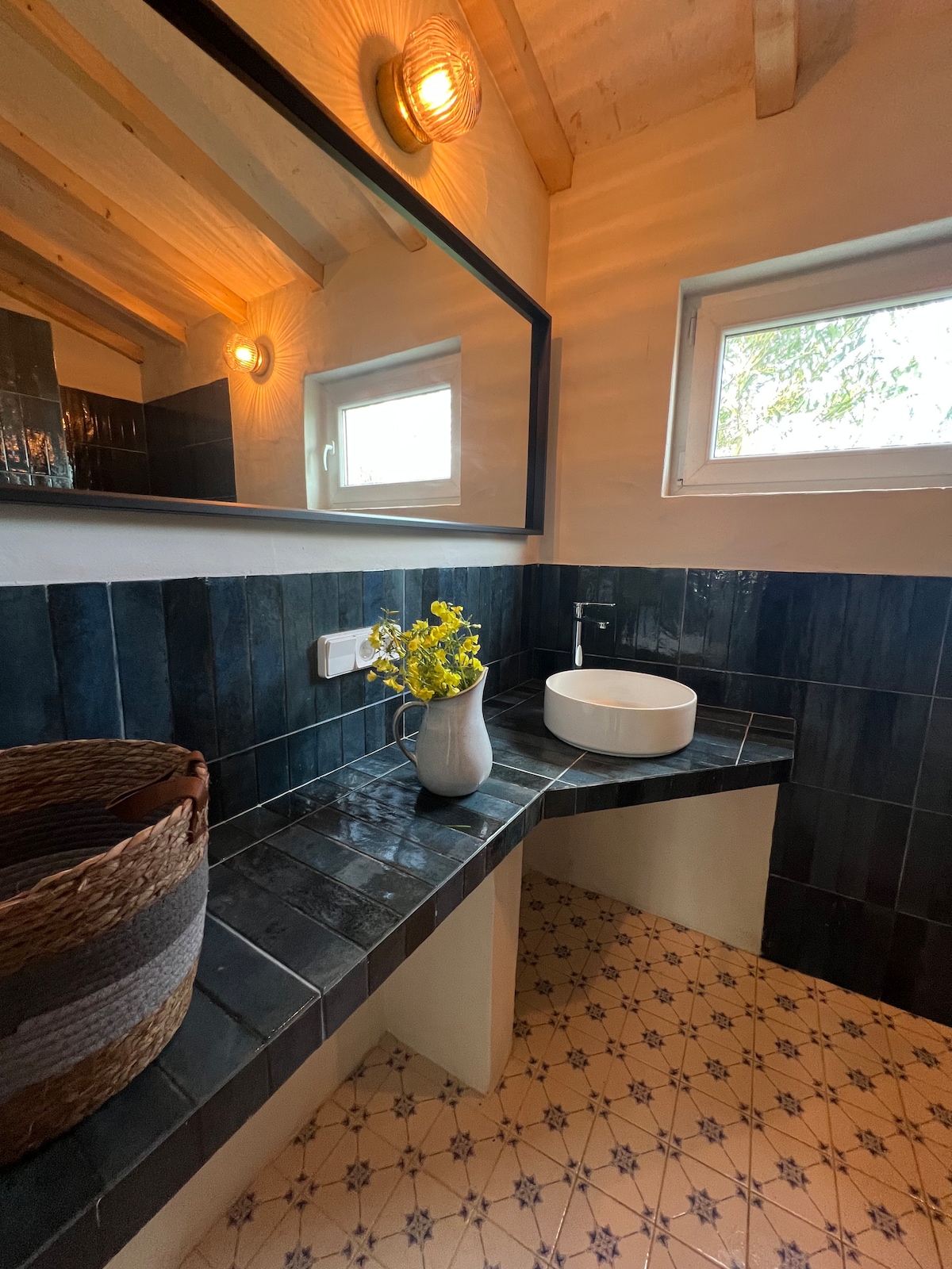The bathroom is equipped with a round vessel sink set against a dark tiled countertop, reflecting a modern design. Natural light enters through a small window, while a mirror above the sink adds depth. A woven basket and a vase of flowers enhance the inviting space.