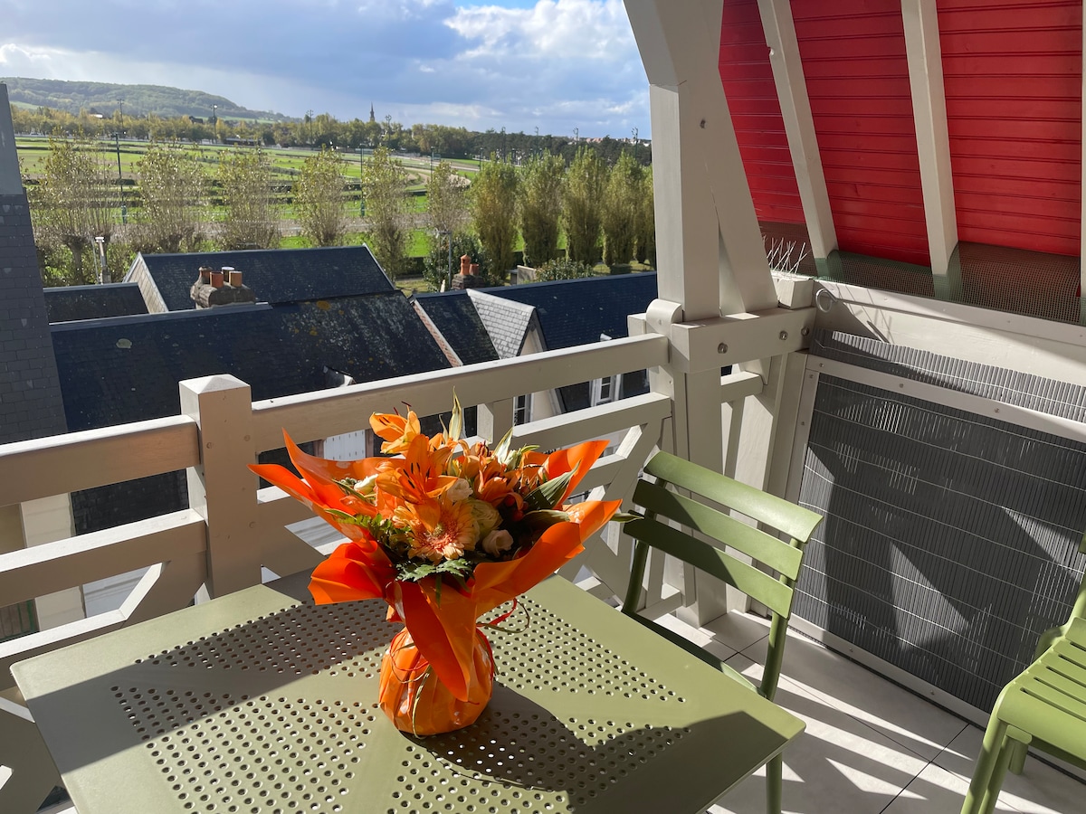 A balcony is featured, adorned with a green table and two matching chairs. A vibrant bouquet of orange flowers sits at the center, contrasting the surrounding landscape of greenery and rooftops. A serene view of fields and distant hills is visible in the background.