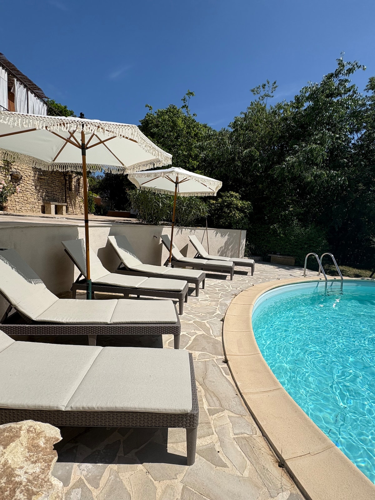 A well-maintained pool area features several lounge chairs arranged beside a sparkling blue pool. Each chair is shaded by a large parasol, providing comfort for relaxation. Lush greenery surrounds the pool, enhancing the serene atmosphere of this outdoor space.