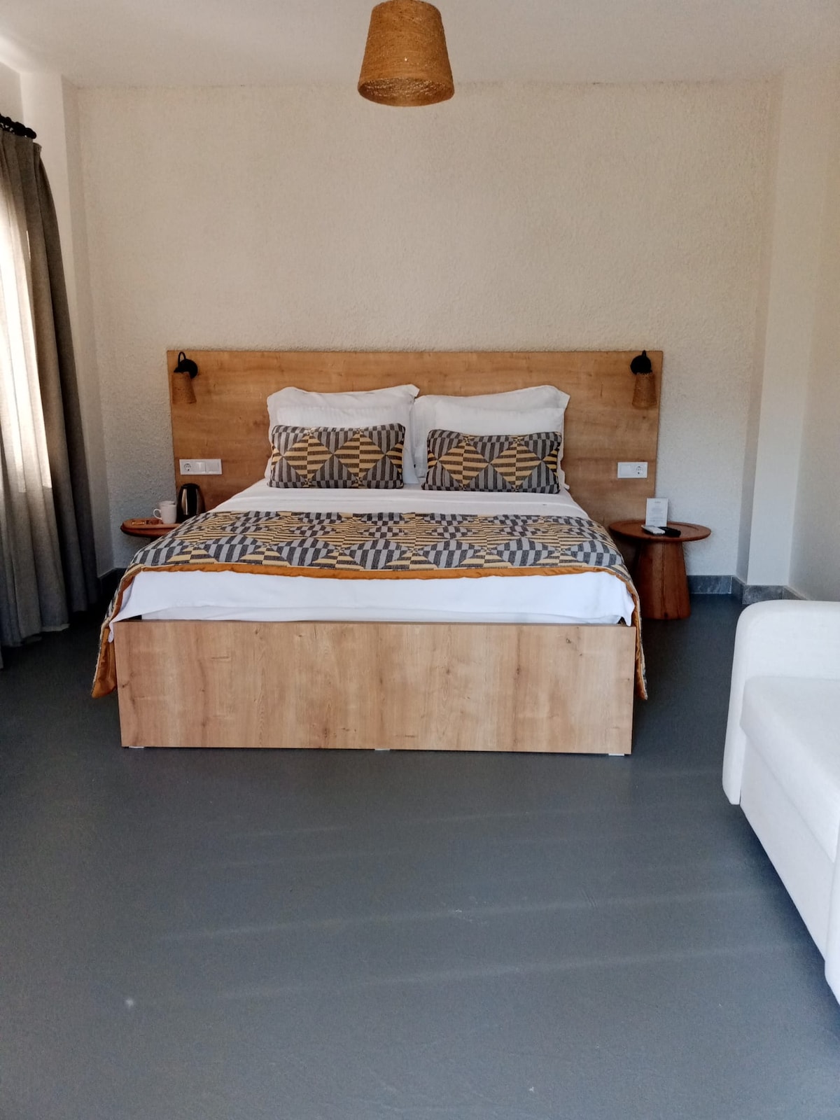 A spacious bedroom is furnished with a king-size bed, dressed in patterned linens. A cozy seating area is provided with a light-colored armchair. Natural light streams in from a nearby window, complemented by soft curtains, and a woven light fixture hangs from the ceiling.