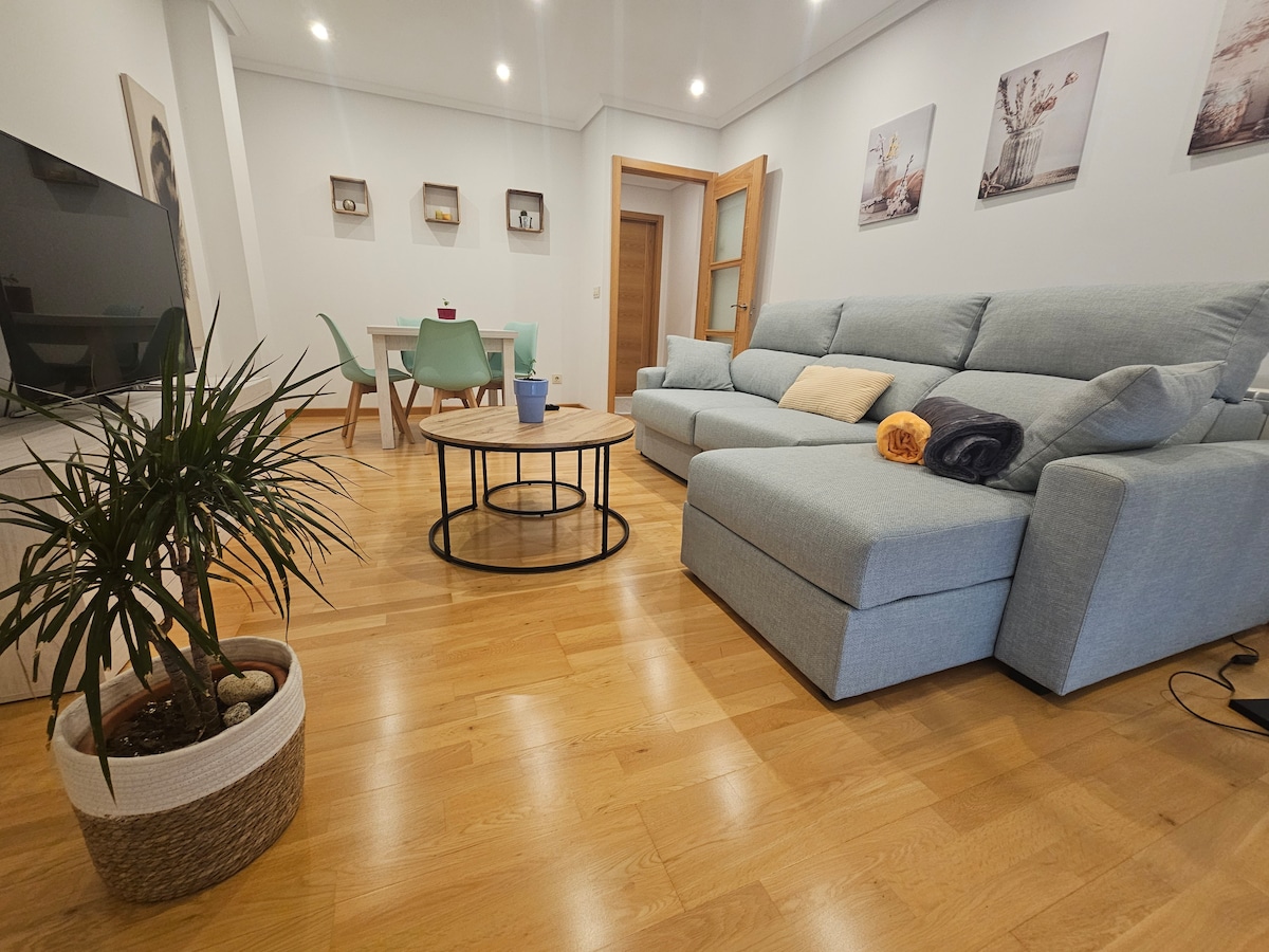 A modern living area features a light blue sectional sofa paired with a round coffee table. A small potted plant adds a touch of greenery to the warm wooden flooring. Nearby, a dining table with colorful chairs is visible, complemented by decorative wall art.