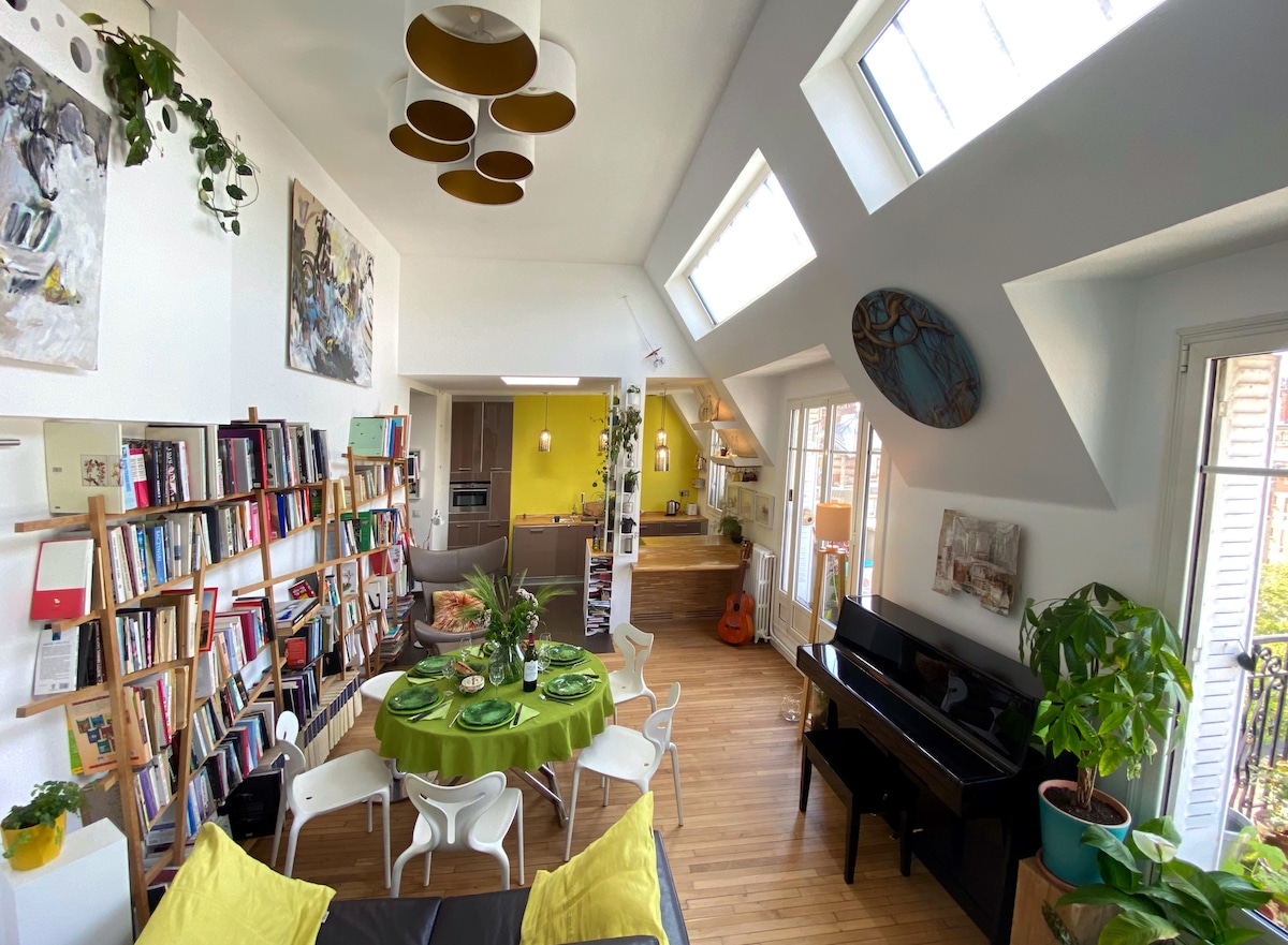 The open living space features a round dining table set with green tablecloth and plants. A wooden bookshelf is filled with books, and a leather sofa is positioned nearby. Large skylights provide ample natural light, illuminating the colorful artwork and plants throughout the room.