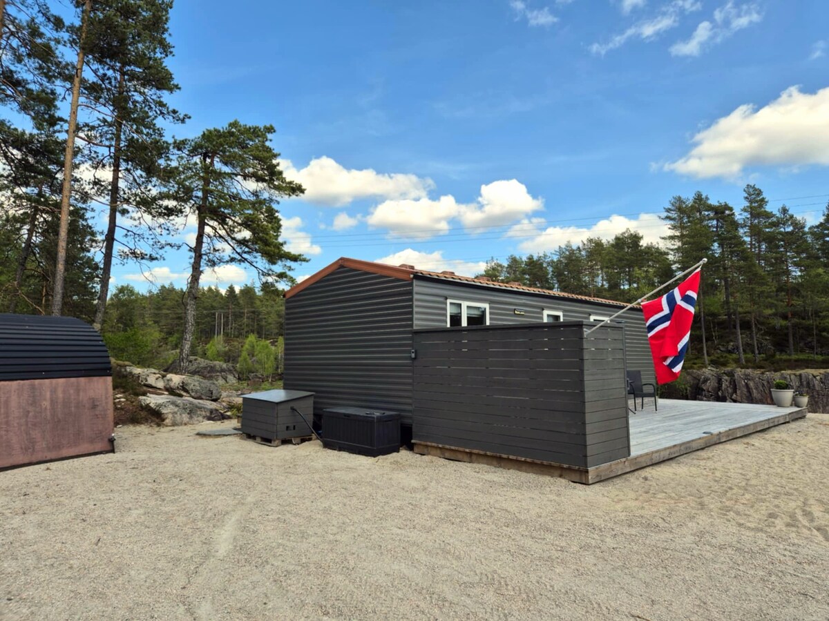 A modern cabin is surrounded by natural landscapes, featuring an outdoor deck with a Norwegian flag proudly displayed. The facade showcases a combination of dark siding and large windows, creating an inviting atmosphere. The sandy area adds to the rustic charm of the setting.