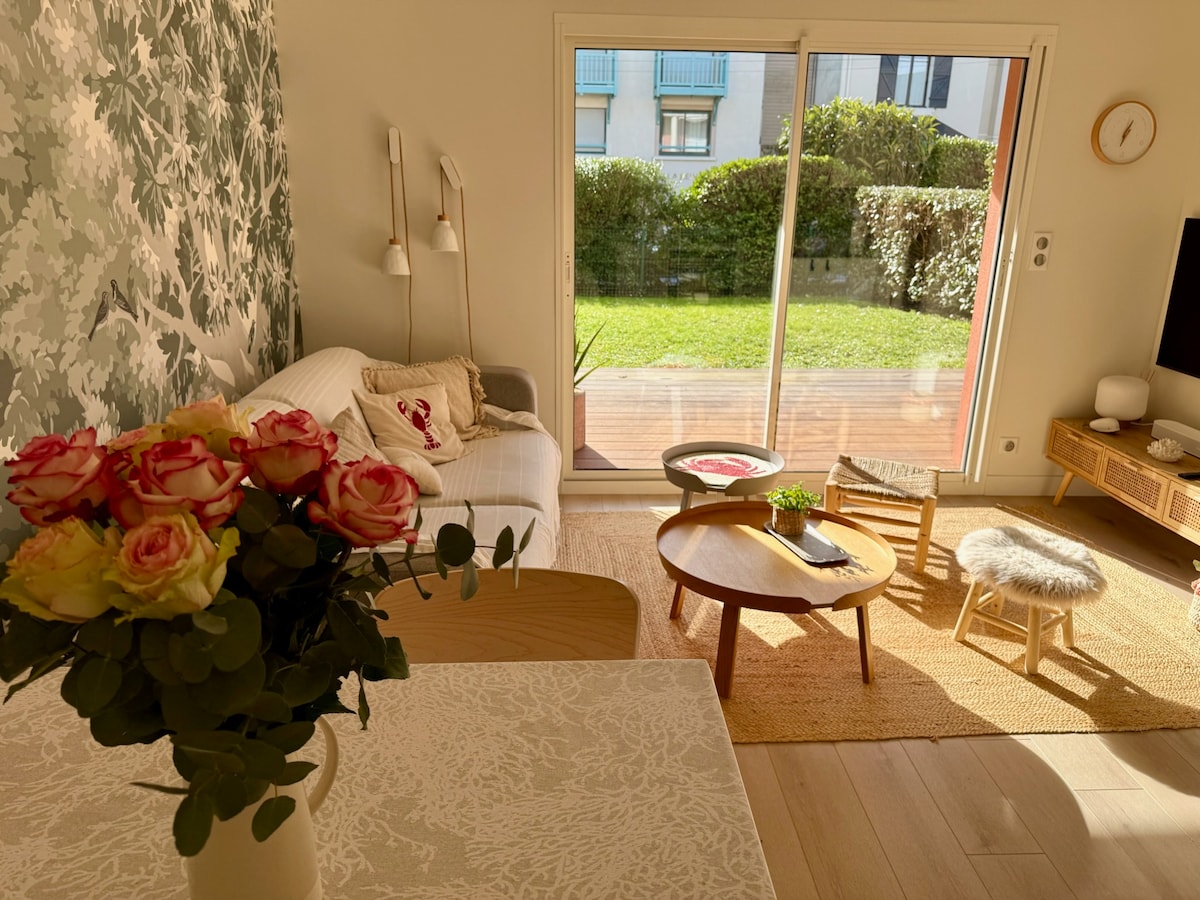 A cozy living area is showcased with a comfortable sofa, accentuated by a floral wall design. Large glass doors offer a view of a lush garden. A decorative vase with fresh roses is placed on a wooden table, accompanied by a small round table and cozy stools.
