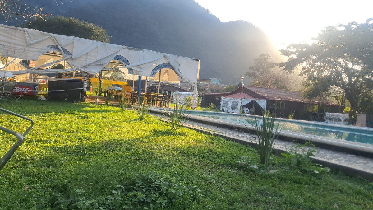 A serene garden area is showcased, featuring lush green grass and a well-maintained swimming pool. Elegant seating arrangements are placed under shaded awnings, while distant hills create a tranquil backdrop. Soft light filters through trees, enhancing the peaceful ambiance of the space.