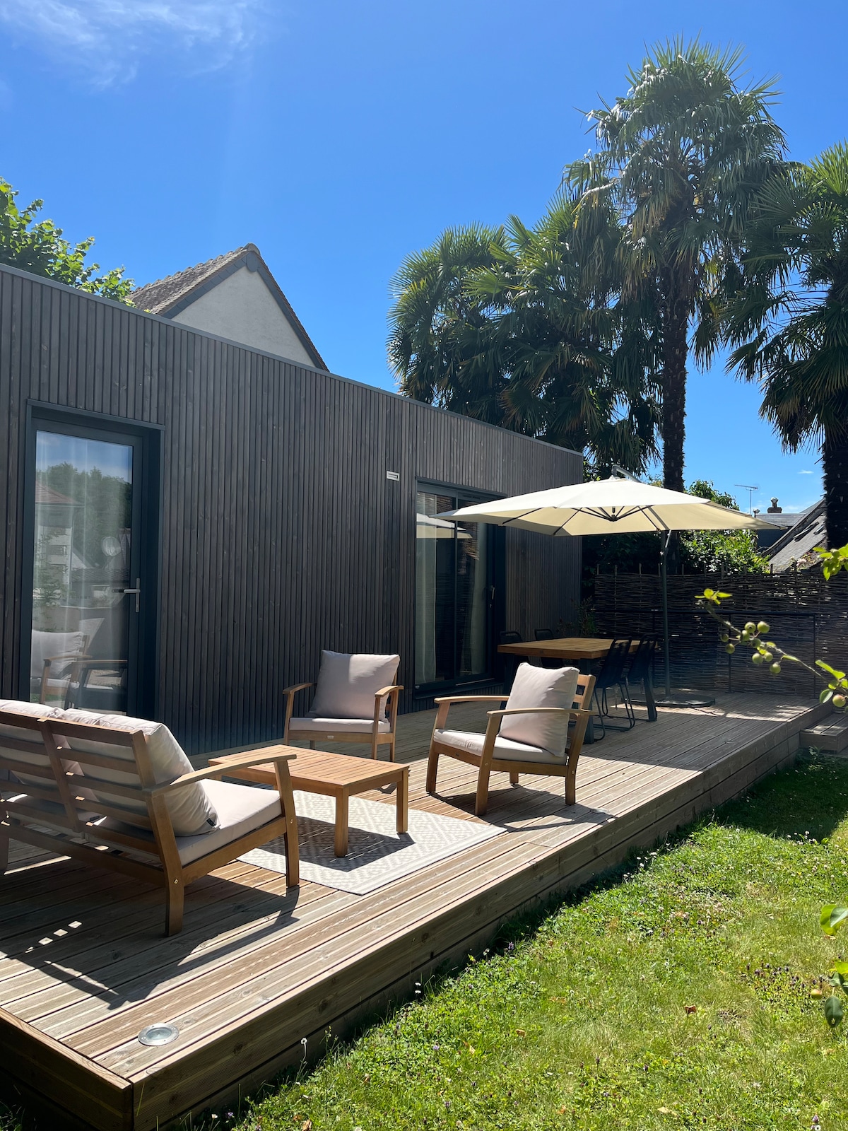 A wooden terrace is furnished with two stylish armchairs and a dining table, set beneath a large umbrella for shade. Lush greenery surrounds the space, while sunlight enhances the natural wooden tones of the exterior.