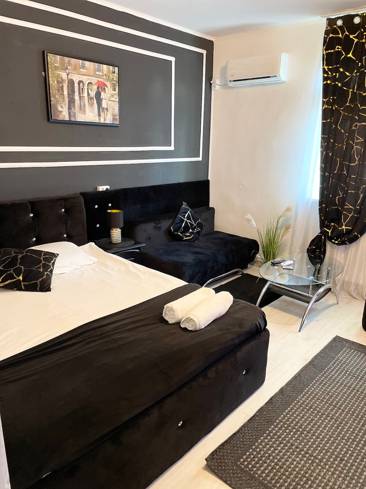 A compact bedroom features a spacious bed with a white linen cover and decorative pillows. A small glass table is positioned beside the bed, accompanied by a stylish rug. A painting hangs above the bed, and natural light enters through a nearby window adorned with dark curtains.