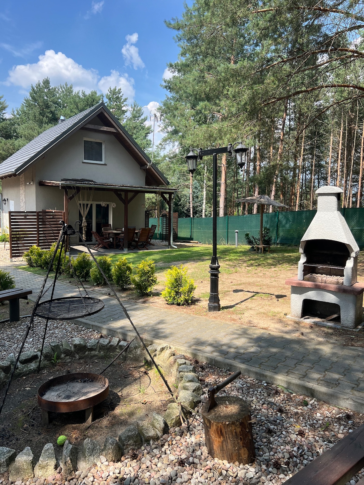 A charming outdoor area features a stone grill and fire pit surrounded by greenery. The house, set back among tall trees, includes a covered terrace with a dining area. Pathways and decorative lanterns enhance the inviting landscape, contributing to a peaceful atmosphere.