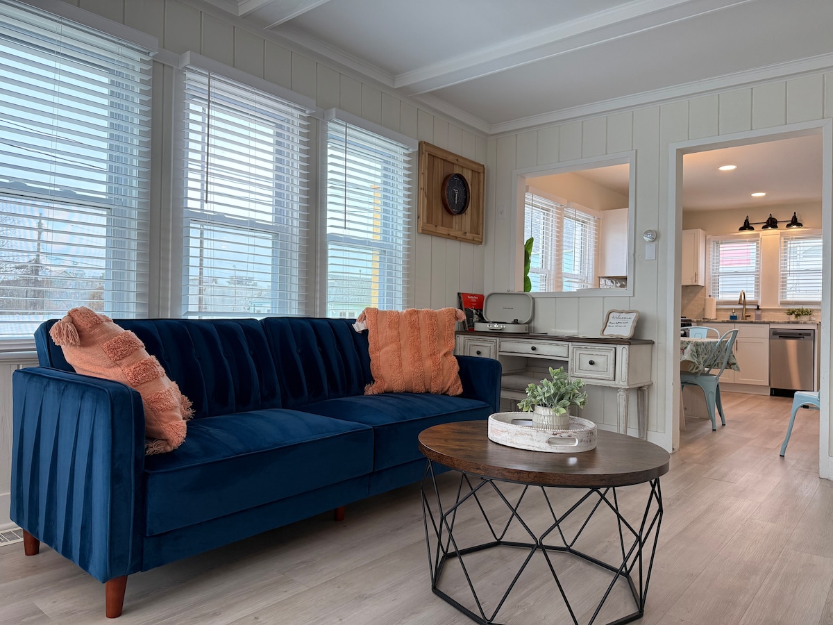 A cozy living area is highlighted by a plush navy blue couch adorned with soft pillows. A round coffee table with a modern design sits in the center, complementing the light-colored walls and bright windows that allow natural light to fill the space.