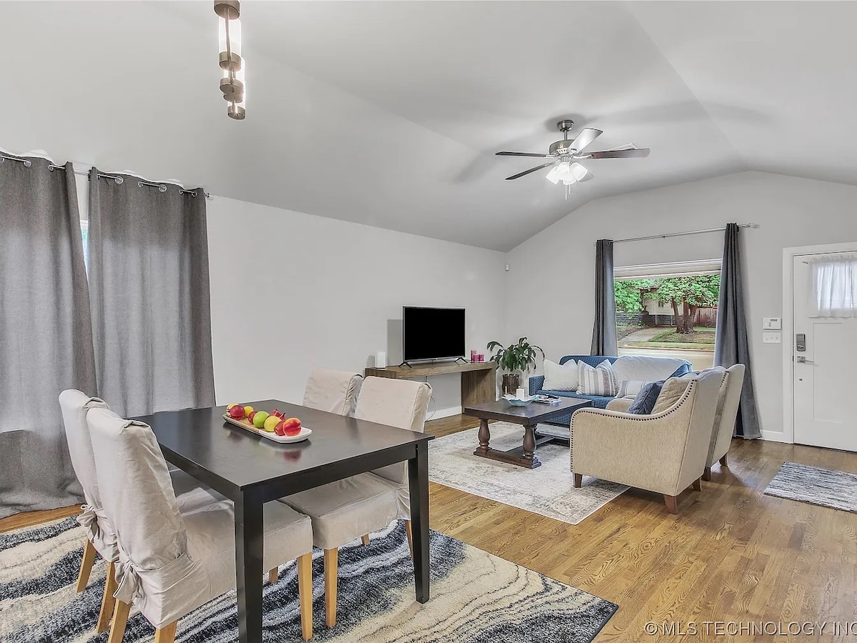 An open living area is featured with a dining table set for four and a comfortable seating arrangement. A television sits on a console, while a plant adds a touch of greenery. Soft natural light filters through large windows adorned with gray curtains.