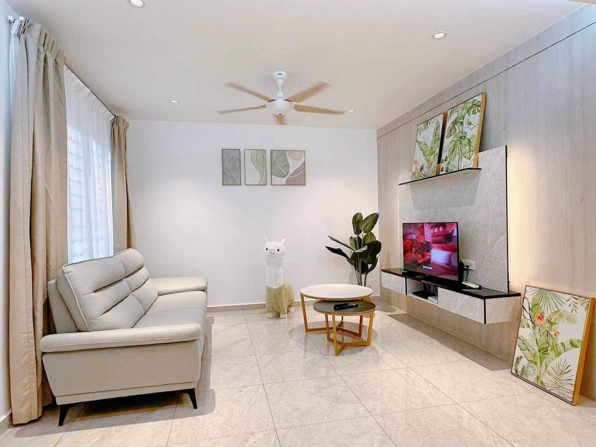 A modern living area is presented with a light-colored sofa and a circular coffee table. A television is mounted on the wall, accompanied by decorative wall art. Natural light filters through sheer curtains, and a potted plant adds a touch of greenery to the space.
