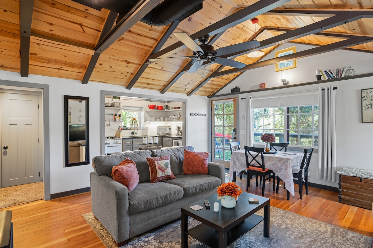 The open living space showcases vaulted wooden ceilings and large windows allowing natural light. A comfortable sofa with decorative pillows faces a dark coffee table. A dining area with a white table and chairs is visible, along with a well-equipped kitchen in the background.
