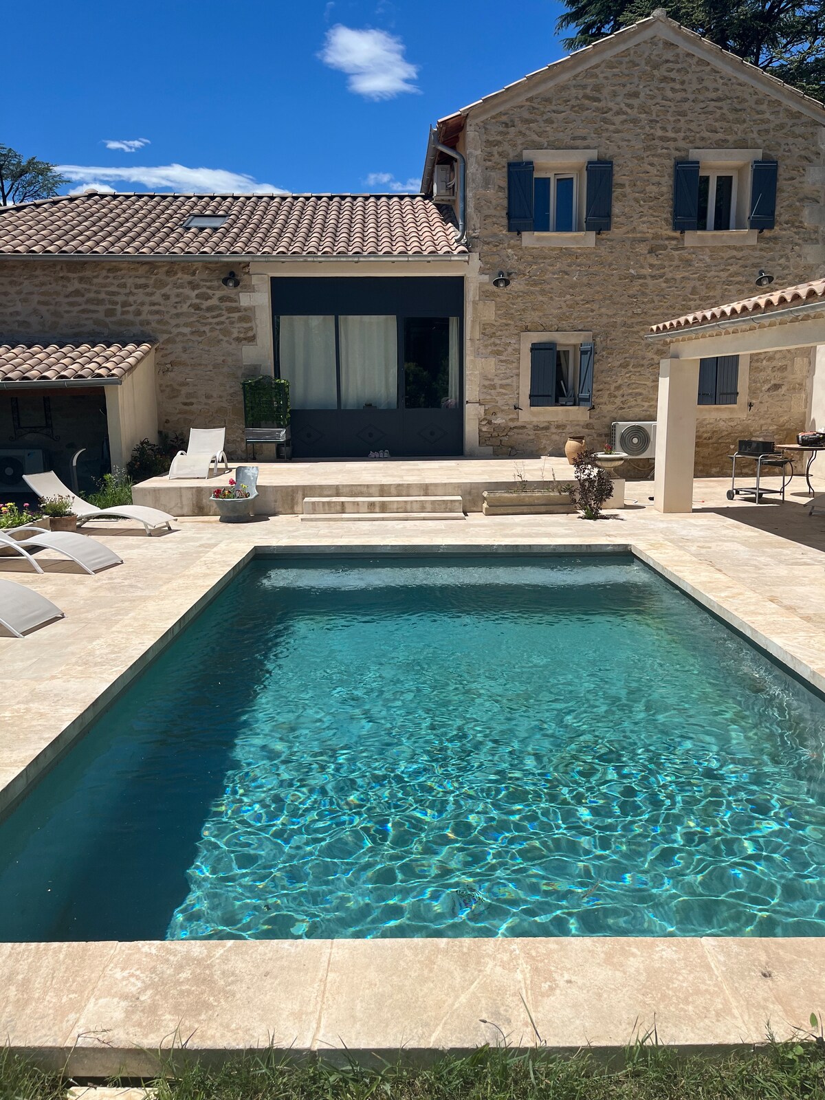 A refreshing pool stretches across the foreground, featuring clear, blue water that reflects sunlight. In the background, a charming stone house is visible, complete with blue-shuttered windows and a tiled roof, surrounded by a well-manicured outdoor space.