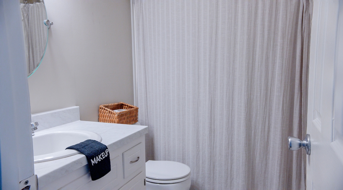 A bathroom is equipped with modern fixtures, featuring a marble countertop and a circular mirror. A white curtain provides privacy for the shower area. Nearby, a wicker basket adds a touch of organization next to the toilet, creating a tidy and functional space.