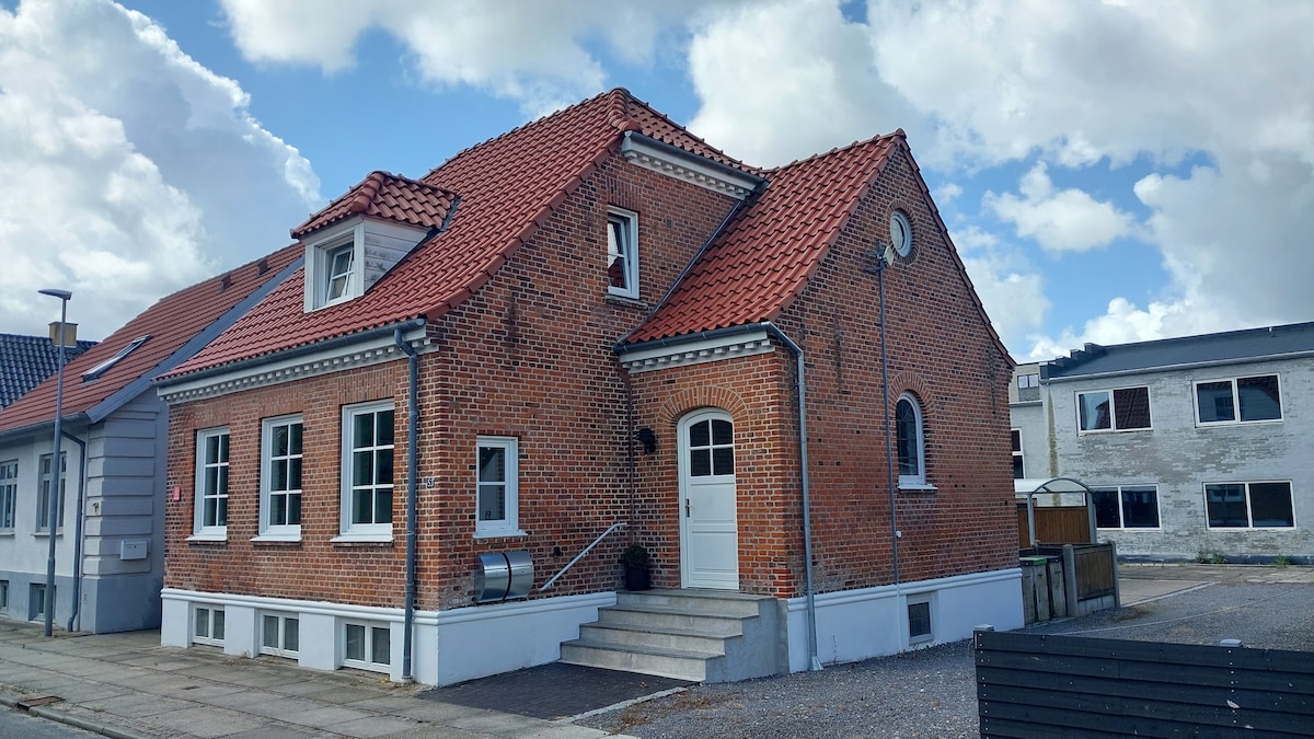 The exterior of the renovated brick house is showcased, featuring a steep roof with bright red tiles. Large windows allow natural light to enter, while a staircase leads to the front door. The surrounding area includes gravel pathways and nearby buildings.