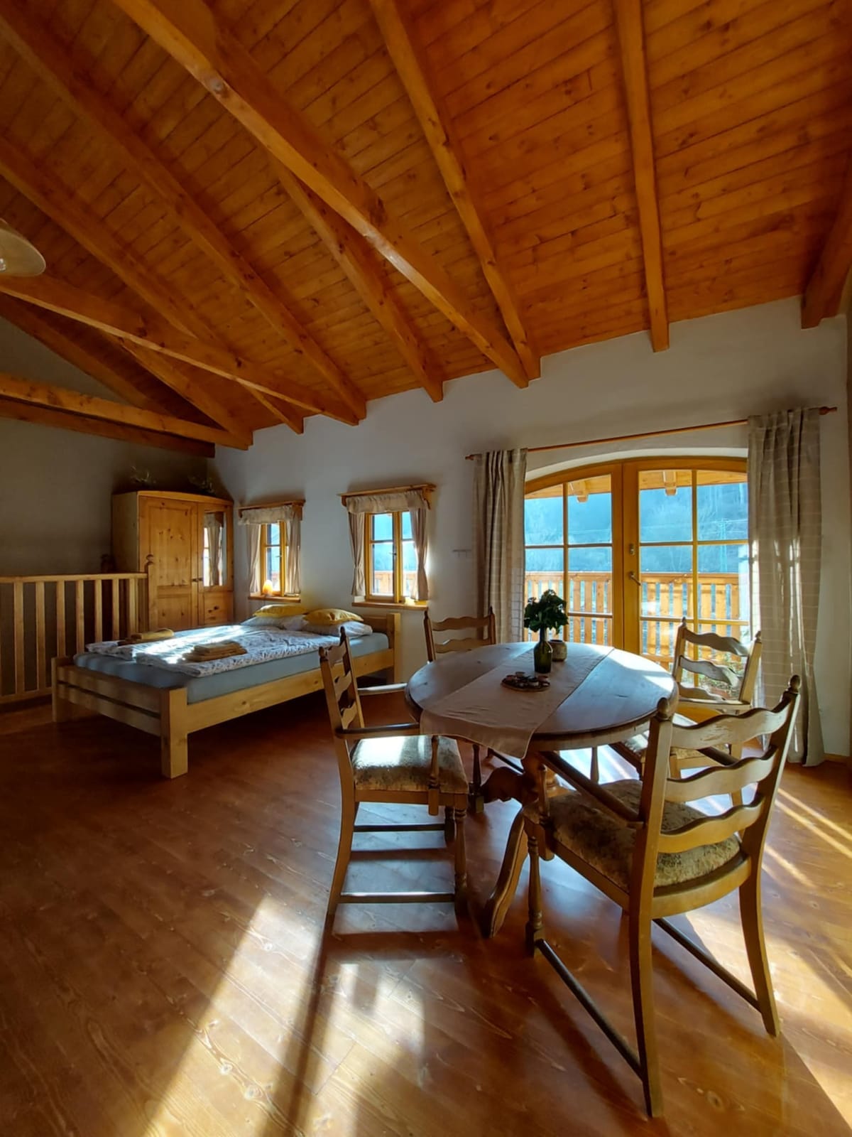 A bright room features a spacious bed with light bedding and natural wood accents. A circular dining table surrounded by wooden chairs is positioned near large windows that open to a balcony, allowing natural light to fill the space.