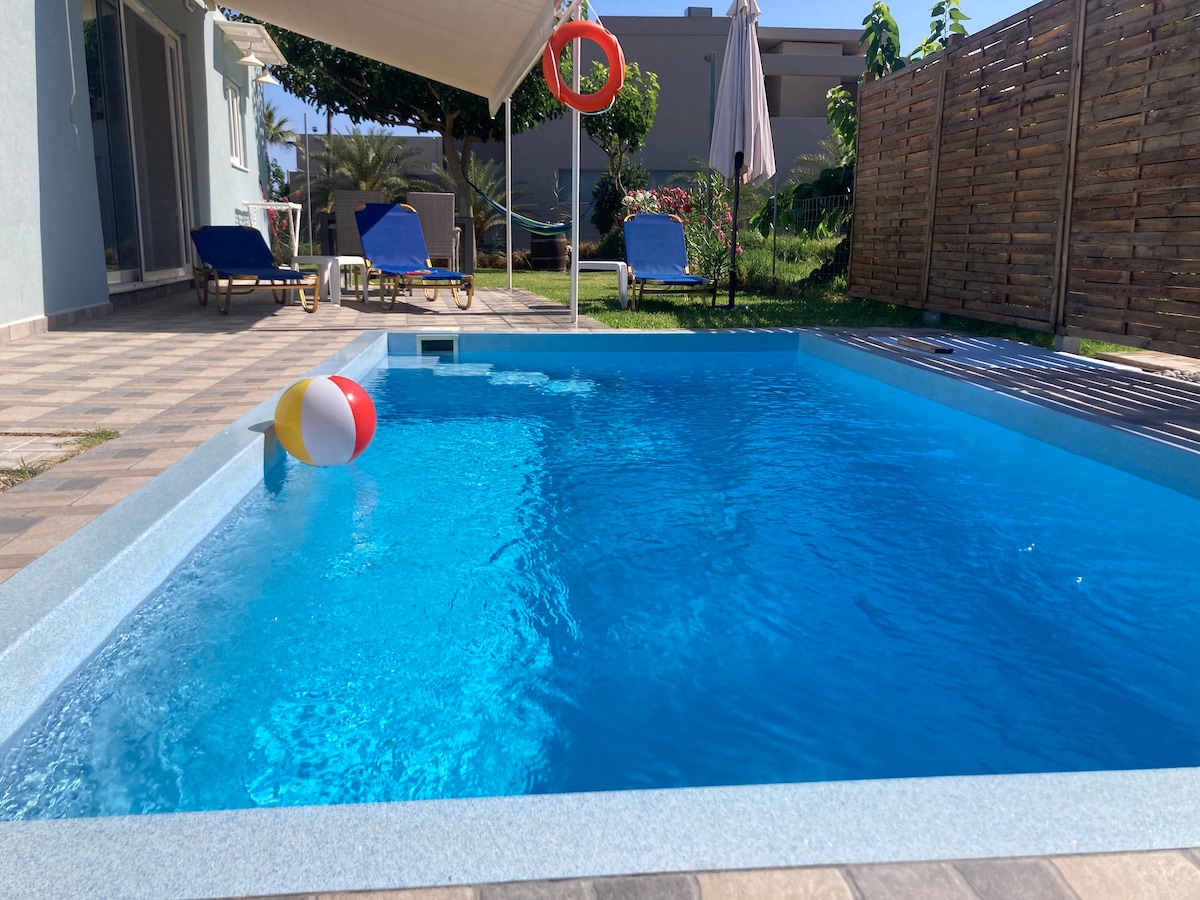 A private swimming pool is seen, featuring clear blue water and a colorful beach ball floating on the surface. Sun loungers are positioned nearby on the patio, with a shaded area created by an awning, and lush greenery surrounds the space.
