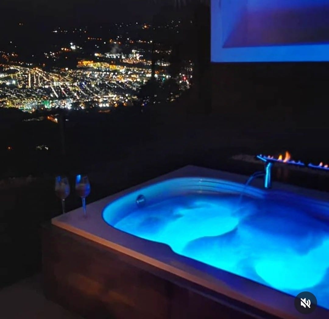A hot tub is illuminated with soft blue light, positioned to overlook a panoramic night view of a sparkling city below. Two glasses are placed nearby, adding to the serene ambiance. The setting invites relaxation while enjoying the surrounding landscape.